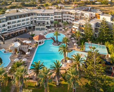 Aerial view of a luxury Greek island resort featuring turquoise swimming pools, palm trees, and modern hotel suites.