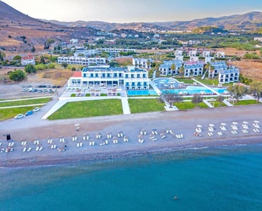 Aerial view of a luxury beachfront resort in Greece with outdoor swimming pools and a pebble beach.