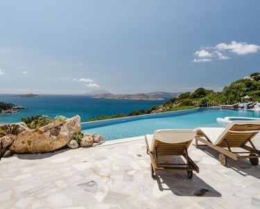 Luxury infinity pool overlooking the Aegean Sea with wooden lounge chairs on a stone terrace.