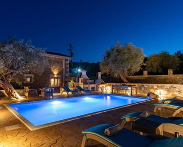 Luxury villa stone patio and private swimming pool illuminated at night with sun loungers.