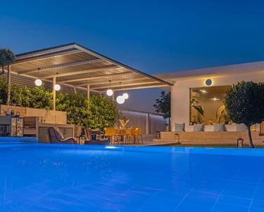 Luxury modern villa patio with blue pool, outdoor kitchen, and illuminated dining area at night.
