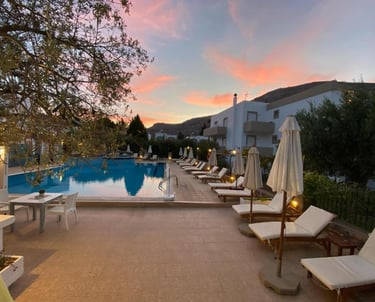 Luxury resort swimming pool with lounge chairs and umbrellas at sunset in Greece.