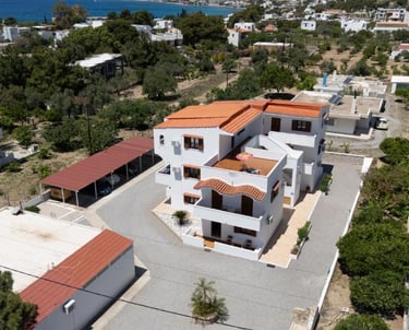 Aerial view of a white multi-story holiday villa with orange roof tiles in a coastal Greek village.