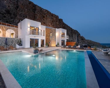 Luxury white villa with illuminated swimming pool at dusk, nestled against a rocky mountain cliff.