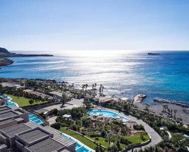 Aerial view of a luxury seaside resort with pools and villas overlooking the blue Aegean Sea.