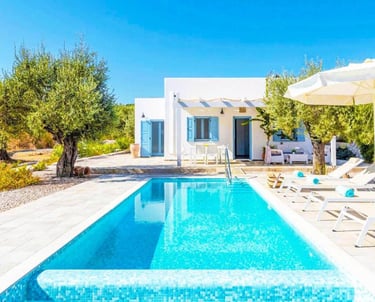 Luxury white Greek villa with a private swimming pool, sun loungers, and olive trees under a clear blue sky.