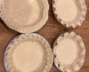 Four unbaked pie crusts with crimped edges in aluminum tins on a wooden surface.