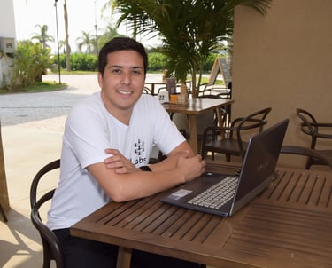 Douglas da Parceria Labs, sorrindo, sentado com notebook sob uma mesa de madeira em um café