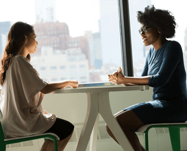 Two professional women sit talking at a workspace table in an office environment.