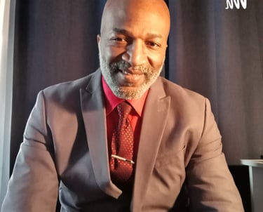 A smiling man posing in a sitting position wearing a grey suit and red tie 