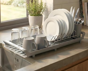 Gray plastic dish drying rack with a self-draining spout holding clean plates, mugs, and glasses on a kitchen counter.
