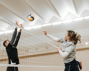 A volleyball player in a gray shirt jumps to spike the ball while an opponent in black reaches up to