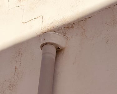 A vertical drainage pipe attached to a white exterior wall with visible cracks and shadows.
