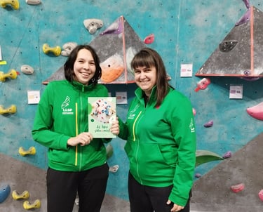 two women in green jackets standing next to a climbing wall holding a book