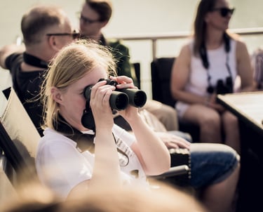 A blonde woman uses black binoculars to look at a distance during a sunny boat tour with group.