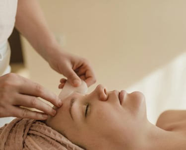 Mujer recibiendo masaje facial con piedra Gua Sha, relajada y con toalla en la cabeza en un ambiente