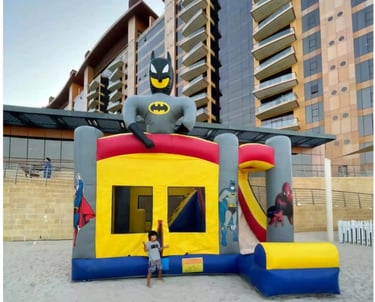 Superhero themed inflatable bounce house with Batman and a slide on a sandy beach.
