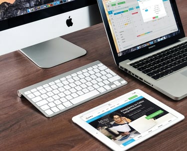 A modern office workspace with an Apple iMac, MacBook laptop, wireless keyboard, and iPad on a wooden desk.