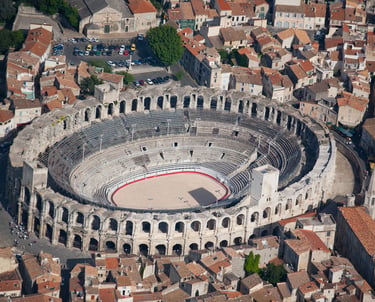 High-angle view of the ancient Roman amphitheater in Arles, France, surrounded by historic city buildings under a bright sky.