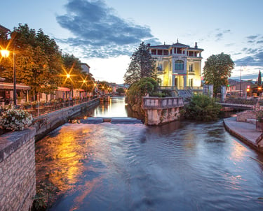 Evening view of L'Isle-sur-la-Sorgue canal in Provence with illuminated riverside cafes and a grand historic building.