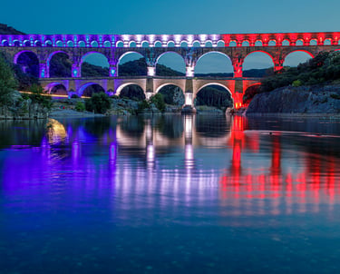 Pont du Gard Roman aqueduct illuminated with blue, white, and red lights reflected in the Gardon River at night.