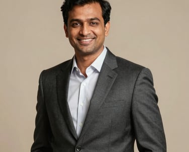 A professional corporate headshot of a South Asian man in his late 30s, wearing a tailored charcoal suit, smiling confidently, wealth consultant style, warm studio lighting.