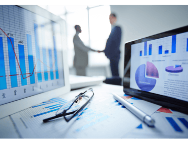 Financial data charts on a digital tablet and monitor with business people shaking hands in the background.