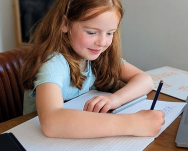 Primary aged girl smiling while writing in a notebook during a tutoring lesson.