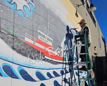Man on a stepladder painting a mural of a tugboat on a wall