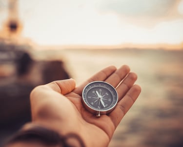 Hand holding a silver pocket compass against a blurred sunset horizon to find his way.