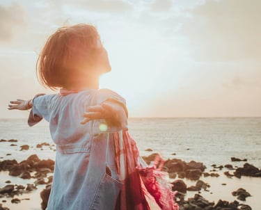 une femme à la retraite, debout, détendue, bras ouverts au bord de la mer, qui inspire le bonheur devant le coucher de soleil