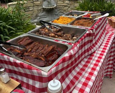 BBQ buffet setup with red checkered tablecloths, ribs, chicken, mac and cheese, and beans