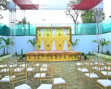 Outdoor wedding stage decoration with yellow fabric, floral backdrop, and gold Tiffany chairs on grass.