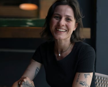 Smiling woman with tattoos sitting at a cafe table, wearing a black shirt and white trousers.