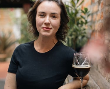 A woman in a black t-shirt sitting at a wooden patio table with a glass of dark beer.