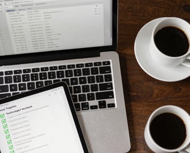 A top-down view of a workspace with a laptop, a tablet, and a cup of coffee. The tablet shows a list of test cases with green checkmarks. Clean and professional.