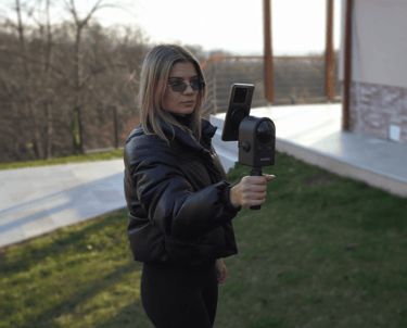 A woman using a handheld 3D scanner and smartphone to capture a digital model outdoors.