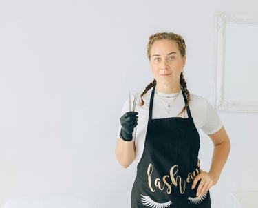 Artista de pestañas profesional con un delantal negro sosteniendo pinzas en un luminoso estudio