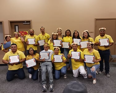 A group of graduates in yellow shirts hold their achievement certificates during a floral crown ceremony.