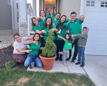 Smiling group in green shirts pointing at a potted boxwood shrub in front of a modern home.