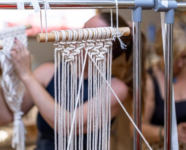 Tejido de macramé en proceso colgado en una barra, con joyera trabajando piezas artesanales al fondo.