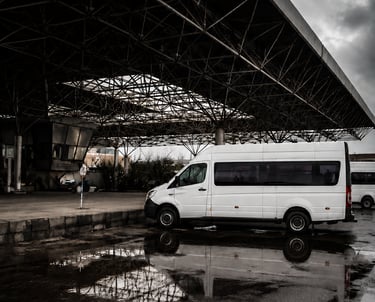 White passenger van parked under a large metal canopy reflected in a rain puddle under a cloudy sky.