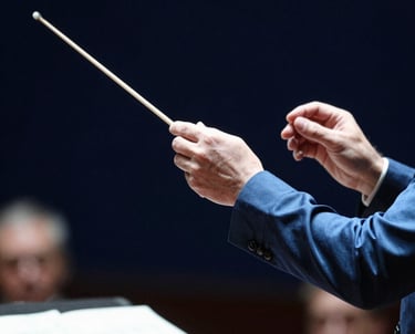 A portrait of a conductor's hand holding a baton, captured mid-motion during a rehearsal. The style is professional and focused, with a blurred dark navy blue background and sharp steel blue highlights on the sleeve.