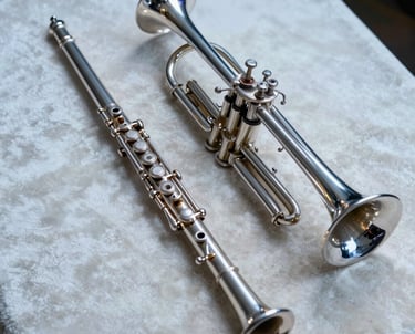 A detailed shot of a flute and a trumpet resting together on a pale off-white velvet cloth. The composition is clean and artistic, using soft steel blue lighting to create an accessible and professional mood.