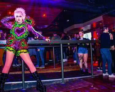 A drag queen in a neon punk outfit performs at a crowded nightclub with vibrant stage lighting.
