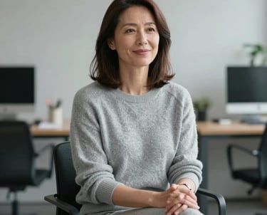 A professional portrait of a female counselor in her 40s, dressed in light grey knitwear, sitting in a minimalist Northern European office with a peaceful expression.