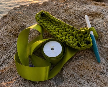 Handmade green crochet ribbon project with a crochet hook resting on seaside rocks at sunset.