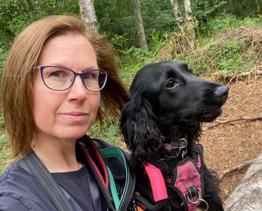 A dog trainer and her black cocker spaniel sit together on a log during a walk