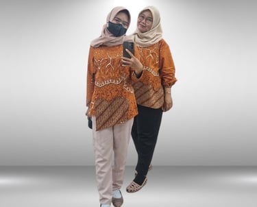 Two women wearing matching orange batik shirts and hijabs posing for a mirror selfie.