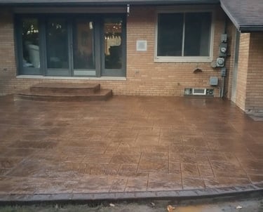Custom brown stamped concrete patio with decorative borders and matching stairs behind a brick house.
